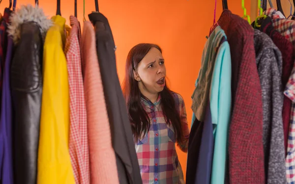 年轻的女人站在衣柜里的衣服中间鬼鬼祟祟的.成年女性选择商店里的东西. — 图库照片