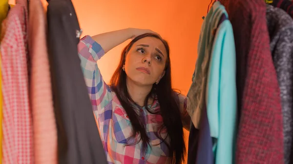 Młoda kobieta grymas stojąc między ubraniami w szafie. Dorosła kobieta wybiera rzeczy w sklepie. — Zdjęcie stockowe