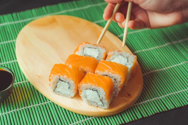 Deliziosi rotoli su tavola di legno su tovagliolo di bambù decorativo. Primo piano di gustosi involtini di sushi con bacchette sul tagliere . — Foto Stock