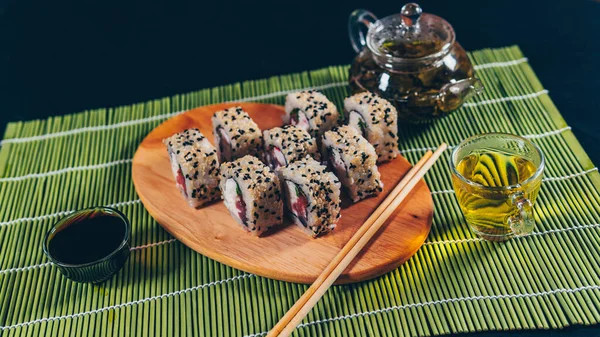 Deliziosi rotoli su tavola di legno con salsa di soia e tè verde su tovagliolo di bambù decorativo. Primo piano di gustosi involtini di sushi con bacchette sul tagliere su sfondo nero . — Foto Stock