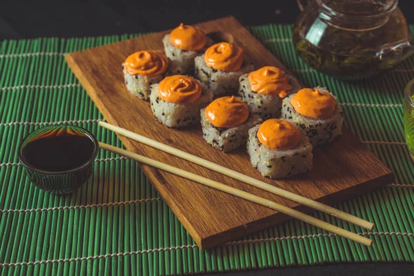 Läckra rullar på träskiva med ätpinnar och grönt te i tekanna på dekorativ bambu servett. Närbild av välsmakande sushi rullar på skärbräda på svart bakgrund. — Stockfoto