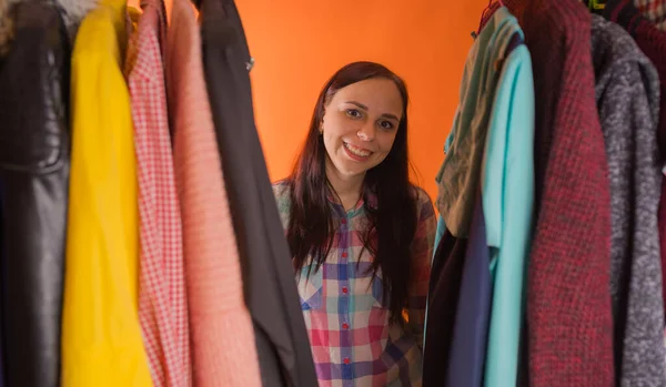 Young woman grimacing standing between clothes in wardrobe. Adult female choosing things in store. Stock Picture