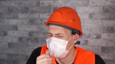 Tulumlu ve maskeli bir erkek inşaat işçisi gri tuğla duvarın arka planında öksürüyor. Enfeksiyon tehdidi kavramı.