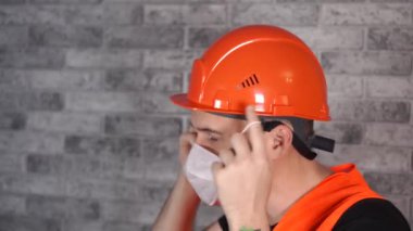 Tulumlu erkek inşaat işçisi, gri tuğla duvarın arka planındaki tıbbi maskeyi çıkarıyor.