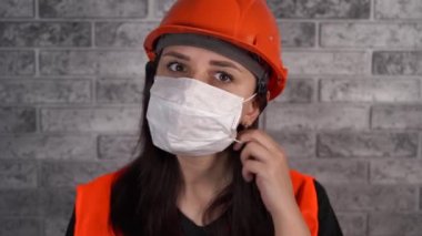Tulumlu kadın inşaat işçisi gri tuğla duvarın arka planındaki tıbbi maskeyi çıkarıyor.