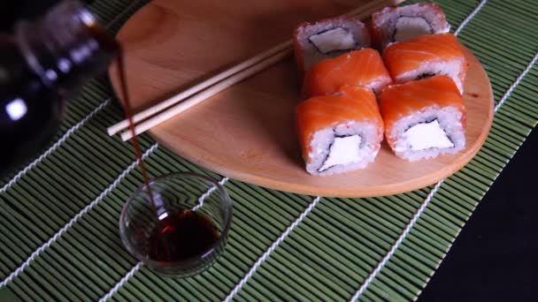 Rolos deliciosos em tábua de madeira com molho de soja em guardanapo de bambu decorativo. Close up de saborosos rolos de sushi com pauzinhos na tábua de corte no fundo preto . — Vídeo de Stock