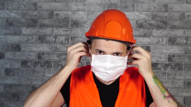 Ouvrier de la construction masculin en salopette portant un masque médical sur le visage sur fond de mur de briques grises . — Video