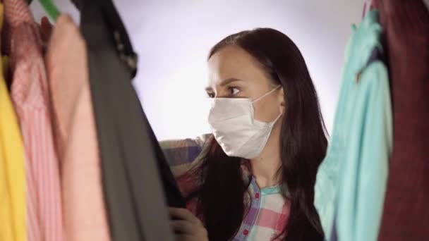 Közelkép fiatal nő orvosi maszk néz körül, gondolkodás, hogy mit válasszon ruhát a szekrényben. — Stock videók