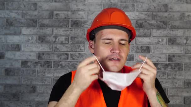 Man bouwvakker in overalls verwijderen van medisch masker uit het gezicht op de achtergrond van grijze bakstenen muur — Stockvideo