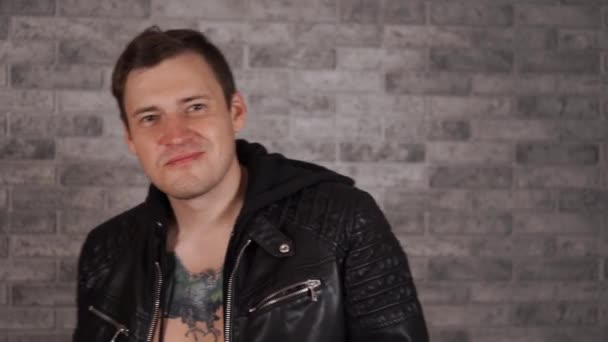 Handsome young man in leather jacket on naked body with tattoo on chest smiling and enticing finger to him. Portrait of brutal charming male, flirting on background brick wall. — Stock Video