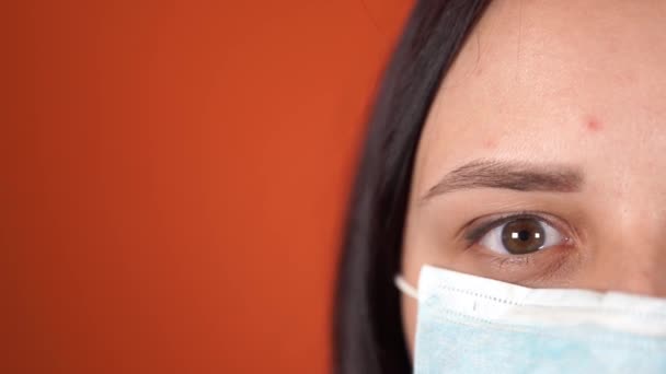 医療マスクをした女性がオレンジの背景に顔を出している。コロナウイルスの流行. — ストック動画