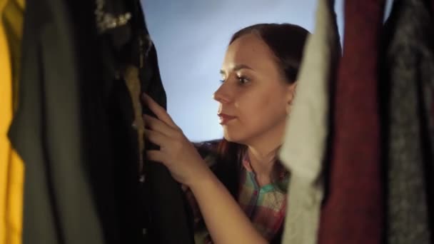 Primer plano de la mujer joven de pie entre la ropa y la elección de las cosas en el armario. Mujer adulta contemplando y buscando qué elegir prendas en tienda . — Vídeos de Stock