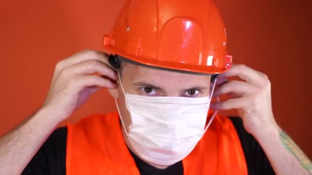 Мужчина строитель в комбинезоне надевает медицинскую маску на лицо на оранжевом фоне . — стоковое видео