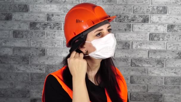 Mujer trabajadora de la construcción en overoles quitando máscara médica de la cara en el fondo de la pared de ladrillo gris — Vídeo de stock