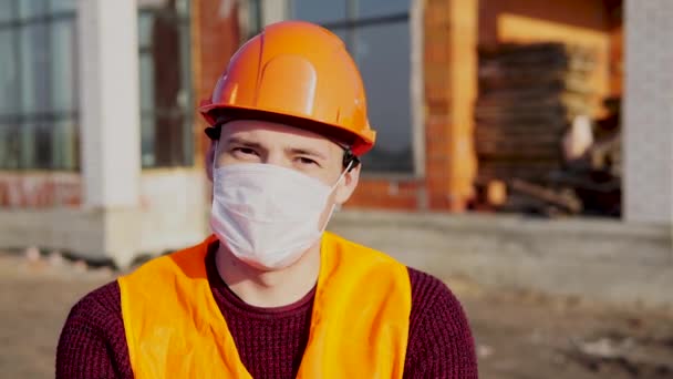 Retrato del trabajador de la construcción masculino en máscara médica y overoles sobre fondo de casa en construcción . — Vídeos de Stock