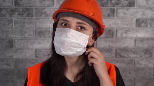 Bauarbeiterin in Overalls entfernt medizinische Maske vom Gesicht auf dem Hintergrund einer grauen Ziegelwand — Stockvideo