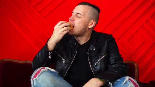 赤い壁に面した小さなソファに腰を下ろしてハンバーガーを食べているハンサムな若い男。大人の男性楽しみます食べ物で彼の足を上にアームチェア. — ストック動画
