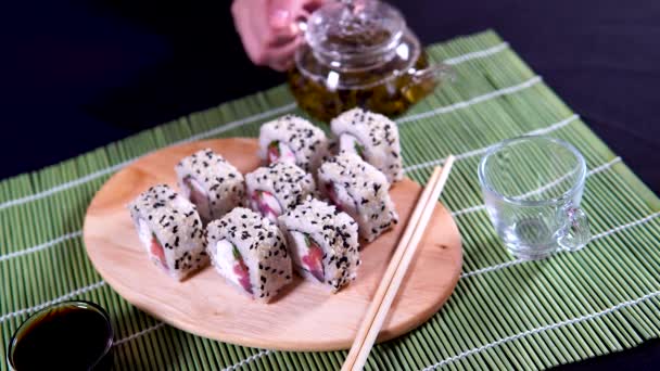 Delicious rolls on wooden board with chopsticks and green tea in teapot on blurry background on decorative bamboo napkin. Close up of tasty sushi rolls on cutting board on black background. — Stock Video