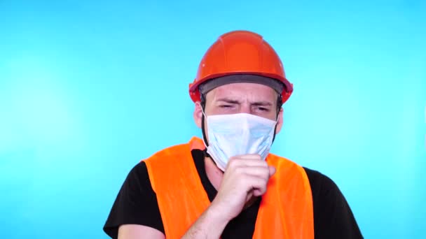 Mannelijke bouwvakker in overall en medisch masker hoesten op blauwe achtergrond. Concept van dreiging van infectie. — Stockvideo