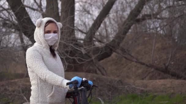 Une jeune femme portant un masque médical et des gants se tient debout avec un vélo, tenant le gouvernail à la campagne. Femme se protégeant contre les maladies à pied. Concept de menace d'infection par le coronavirus . — Video