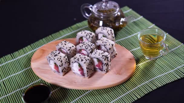 Heerlijke broodjes op houten plank met sojasaus en groene thee op decoratieve bamboe servet. Close-up van lekkere sushi broodjes met eetstokjes op snijplank op zwarte achtergrond. — Stockvideo