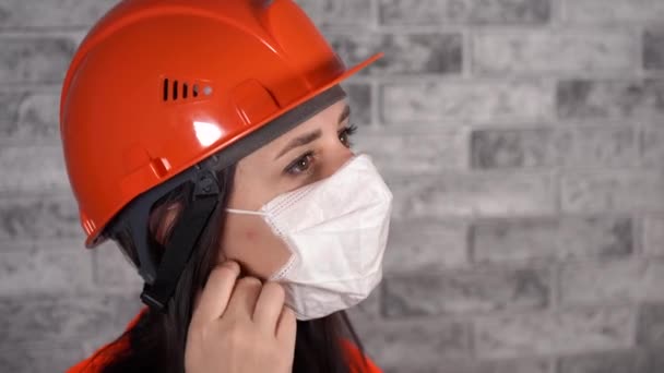 Tulumlu kadın inşaat işçisi gri tuğla duvarın arka planındaki tıbbi maskeyi çıkarıyor. — Stok video