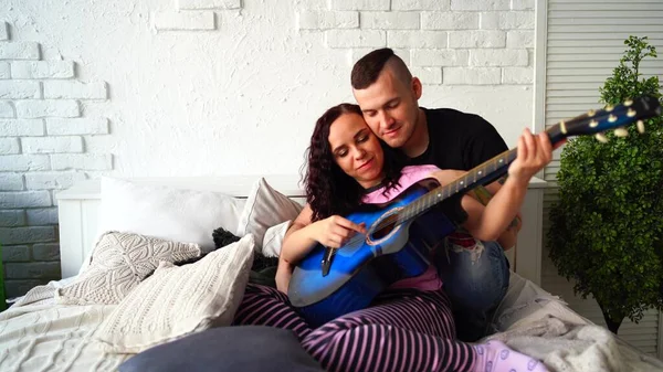 Liebespaar mit Gitarre im Bett. Mann umarmt Freundin, die blaue Akustikgitarre spielt, während sie im Bett sitzt und Zeit zu Hause verbringt — Stockfoto