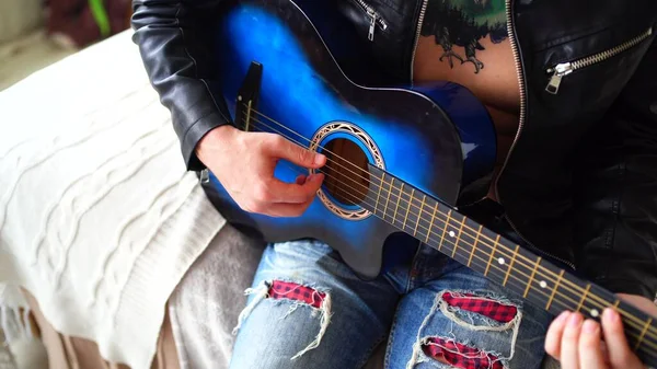 Un joven con un tatuaje en el pecho toca la guitarra en el dormitorio. Hipster aprende a tocar la guitarra. Primer plano de una mano de hombre tocando una cuerda de guitarra. Concepto: músico de rock —  Fotos de Stock