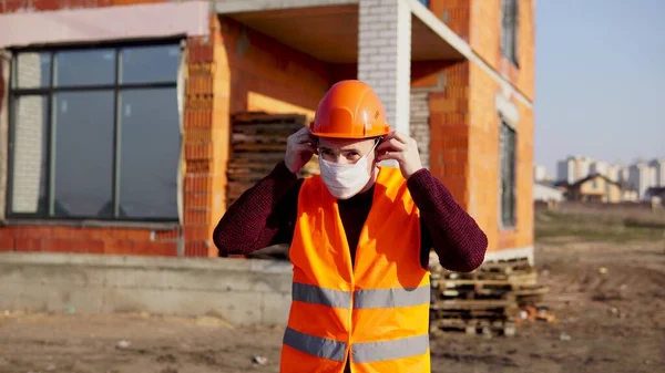 Мужчина строитель в комбинезоне надевает медицинскую маску на лицо на строительной площадке . — стоковое фото