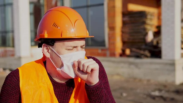 Hombre trabajador de la construcción en overoles y máscara médica tos en el fondo de la casa en construcción. Concepto de amenaza de infección . —  Fotos de Stock