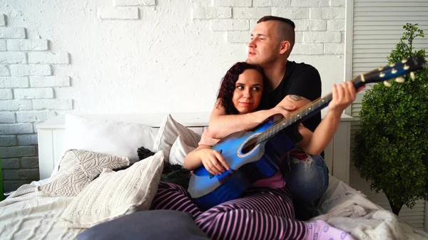 Coppia amorevole con chitarra seduta sul letto. Uomo abbracciare fidanzata giocare chitarra acustica blu mentre seduto sul letto abbracciare e trascorrere del tempo a casa — Foto Stock