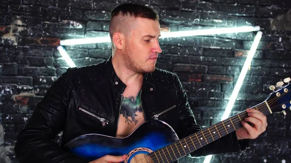 Wytatuowany facet grający na gitarze przy oświetlonej ścianie. Stylowy facet z tatuażem ptaka na klatce piersiowej odwracając wzrok i grając na gitarze, stojąc przed brudną ceglaną ścianą z trójkątnym oświetleniem — Zdjęcie stockowe