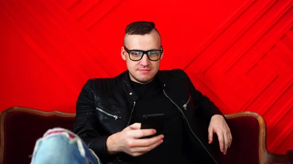 Beau jeune homme avec des lunettes reposant avec smartphone sur un petit canapé contre un mur rouge. Homme adulte assis sur un fauteuil avec téléphone portable dans les mains . — Photo