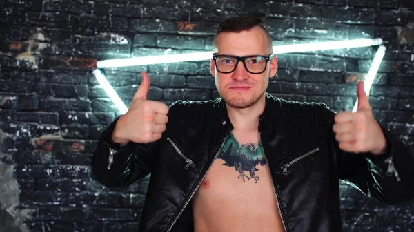 Mannen i trendig jacka tatuering på bröstet stående med en mot upplyst vägg. Ett leende och visar tummen upp. En man visar en gest av gillande. Begreppet gillar eller ogillar. — Stockfoto