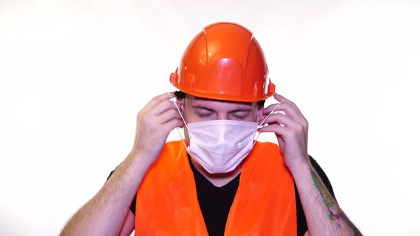 Мужчина строитель в комбинезоне надевает медицинскую маску на лицо на белом фоне . — стоковое фото