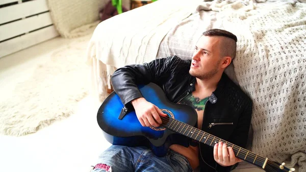 Młody mężczyzna z tatuażem na piersi gra na gitarze w sypialni. Hipster uczy się grać na gitarze. Zbliżenie męskiej dłoni grającej na gitarze. Koncepcja: muzyk rockowy — Zdjęcie stockowe