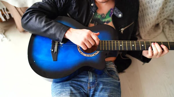 Un giovane uomo con un tatuaggio sul petto suona la chitarra in camera da letto. Hipster impara a suonare la chitarra. Primo piano di un uomo che suona una corda di chitarra. Concetto: musicista rock — Foto Stock