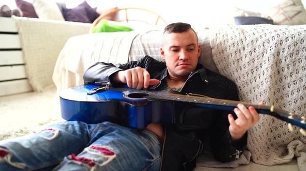 胸にタトゥーのある若い男が寝室でギターを弾いている。ヒップスターはギターを弾くことを学ぶ。ギターの弦を弾く男性の手のクローズアップ。コンセプト:ロックミュージシャン — ストック写真