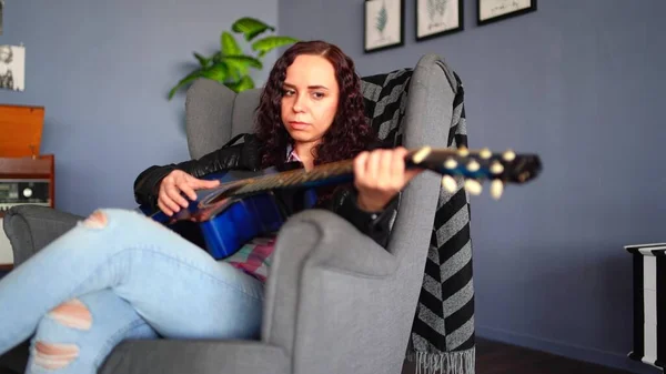 Jonge mooie vrouw in casual kleding zittend in een fauteuil en gitaar spelend thuis. Portret van een mooie vrouw die alleen rust met een muziekinstrument. — Stockfoto