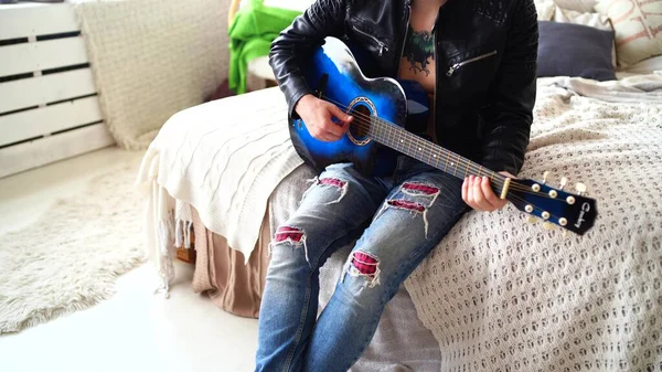 A young man with a tattoo on his chest plays the guitar in the bedroom. Hipster learn to play the guitar. Close-up of a mans hand playing a guitar string. Concept: rock musician — Stock Photo, Image