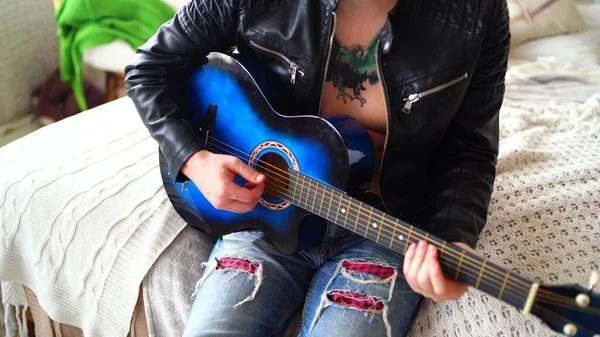 Un giovane uomo con un tatuaggio sul petto suona la chitarra in camera da letto. Hipster impara a suonare la chitarra. Primo piano di un uomo che suona una corda di chitarra. Concetto: musicista rock — Foto Stock