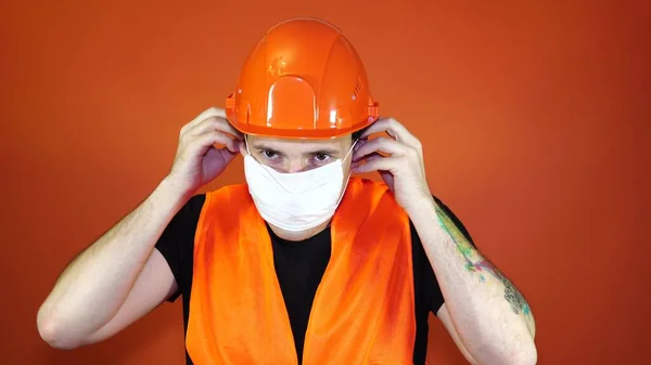Мужчина строитель в комбинезоне надевает медицинскую маску на лицо на оранжевом фоне . — стоковое фото