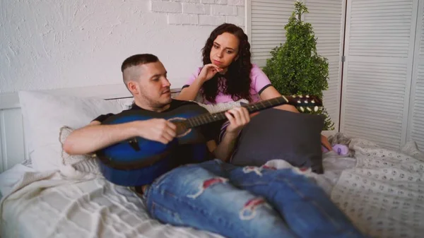 Yatakta oturan gitarlı sevgi dolu bir çift. Mavi akustik gitar çalan bir adam kız arkadaşın için yatakta oturuyor ve evde vakit geçiriyor. — Stok fotoğraf