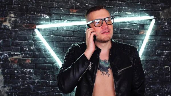 Stylový mladý muž s brýlemi v kožené bundě na nahém těle s tetováním na hrudníku vytáčení telefonu a mluvení na pozadí ošuntělé cihlové zdi s svítící lampy v podobě trojúhelníku. — Stock fotografie