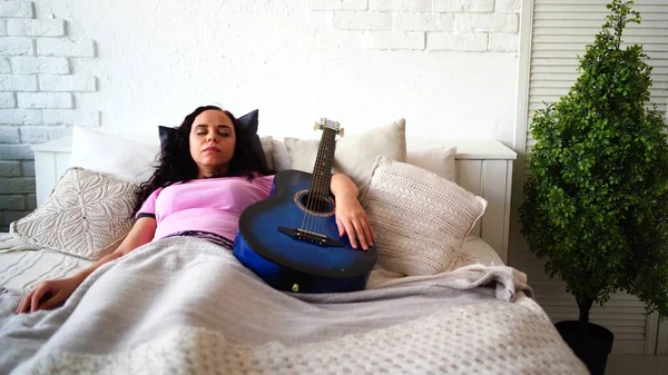 Giovane donna che riposa in abbraccio con la chitarra in camera. Donna adulta in pigiama che dorme con strumento musicale, sdraiata su cuscini sotto il plaid a letto. — Foto Stock