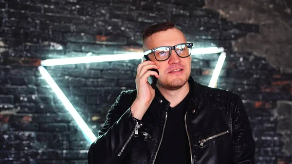 Stylish young man with glasses talking on mobile phone on background shabby brick wall with glowing lamps. Portrait of handsome man speaking on smartphone while standing against illuminated wall.