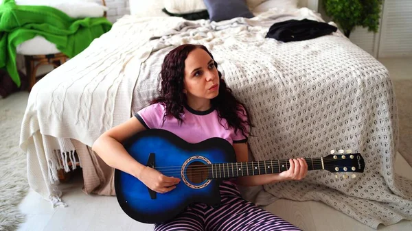 Giovane donna che suona la chitarra in camera da letto. Giovane donna che suona la chitarra nel suo letto. Le piace il suo tempo libero. Bella ragazza suonare la chitarra in una camera da letto accogliente — Foto Stock