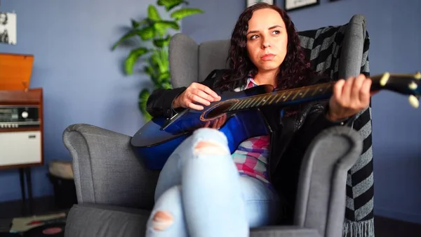 Giovane bella donna in abiti casual seduta in poltrona e suonare la chitarra a casa. Ritratto di bella femmina che riposa sola con lo strumento musicale. — Foto Stock