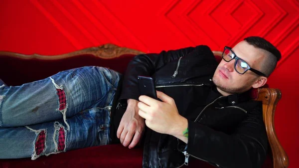 Beau jeune homme avec des lunettes reposant dans un téléphone portable sur un petit canapé contre un mur rouge. adulte charmant mâle retournant à travers diverses nouvelles dans le téléphone mobile, couché sur le côté sur le fauteuil . — Photo
