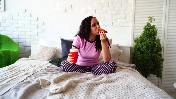 Młoda kobieta w łóżku je hot doga i pije kawę. Śniadanie do łóżka. Dostawa żywności do domu. Kobieta je soczystego hot doga w przytulnej sypialni. Koncepcja: Pokusa na dietę i fast food — Zdjęcie stockowe